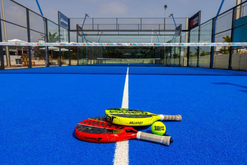 Divi padel raketes ar spilgtām krāsām atrodas uz zilo laukumu, blakus pie tīkla. Fons attēlo modernu sporta laukumu.