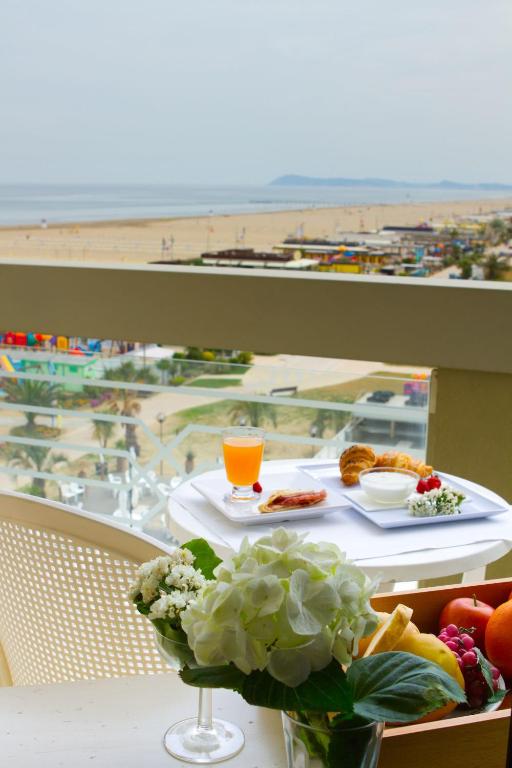 Balkona skats uz pludmali ar brokastu komplektu, kas ietver augļus, konditorejas izstrādājumus un dzērienu. Viegla gaisotne ar jūras ainavu fona.