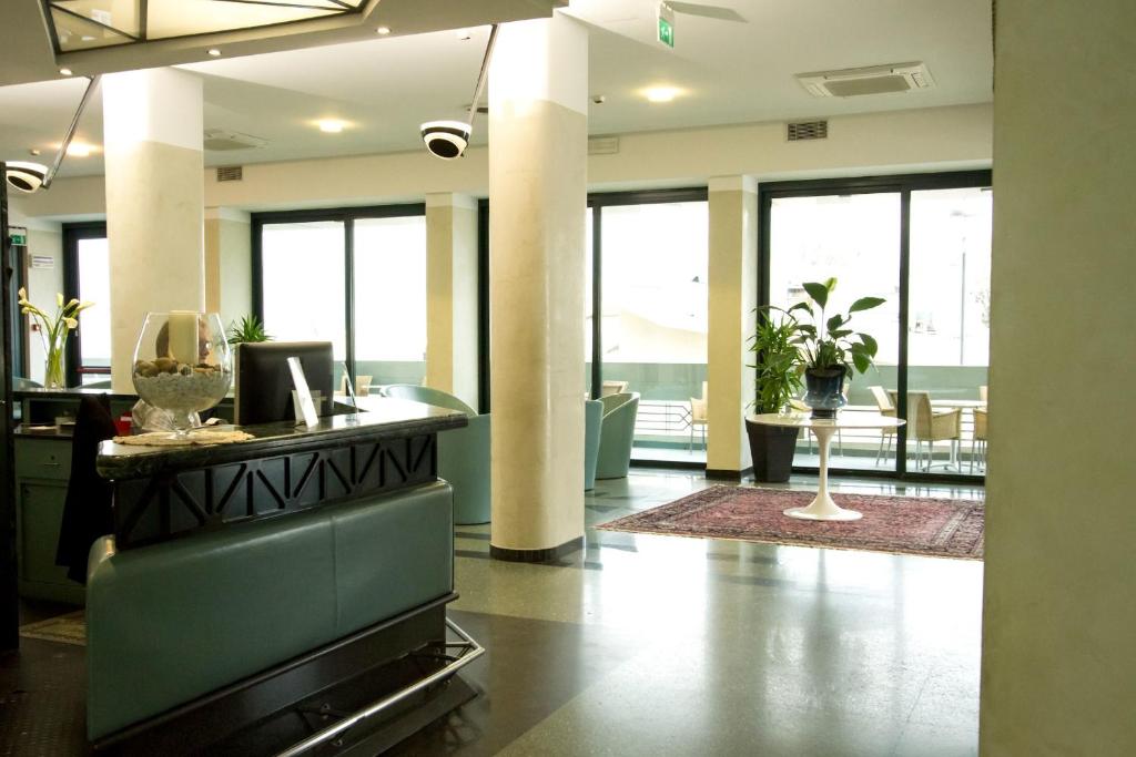 Mūsdienīgs viesnīcas reģistratūras interjers ar plašām logiem un zaļām augiem, radot viesmīlīgu atmosfēru. Stilīga mēbeļu izvēle un gludas līnijas veido elegantu un komfortablu vidi.