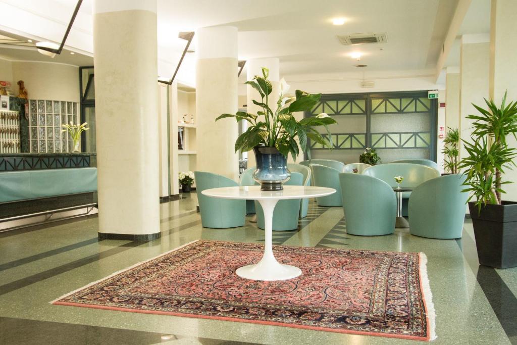 Mūsdienīgs viesnīcas vestibila interjers ar stilīgu galdu, ziedu kompozīciju un ērtām sēdvietām. Telpā ir gaiši toņi un zaļumi, radot relaksējošu atmosfēru.