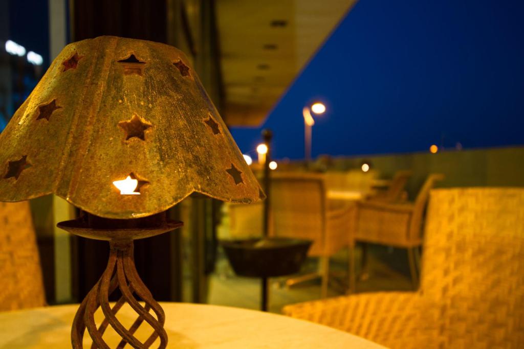 Metāla lampa ar zvaigžņu izgriezumiem, kas rada siltu gaismu, atrodas uz galda. Fons ir nakts ainava ar mēbelēm un gaismas avotiem.