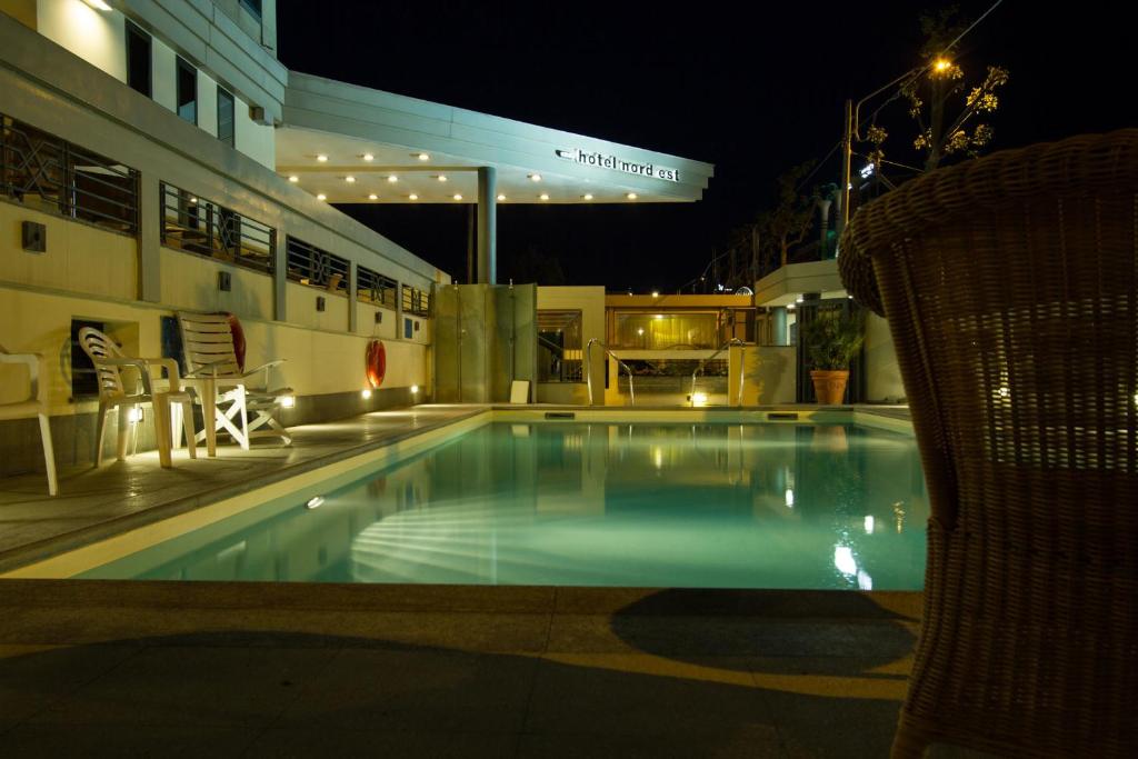 Nakts skats uz viesnīcas baseinu, apgaismotu ar mīkstu gaismu, ar tuvumā esošiem krēsliem un viesnīcas nosaukumu redzamu fona daļā.