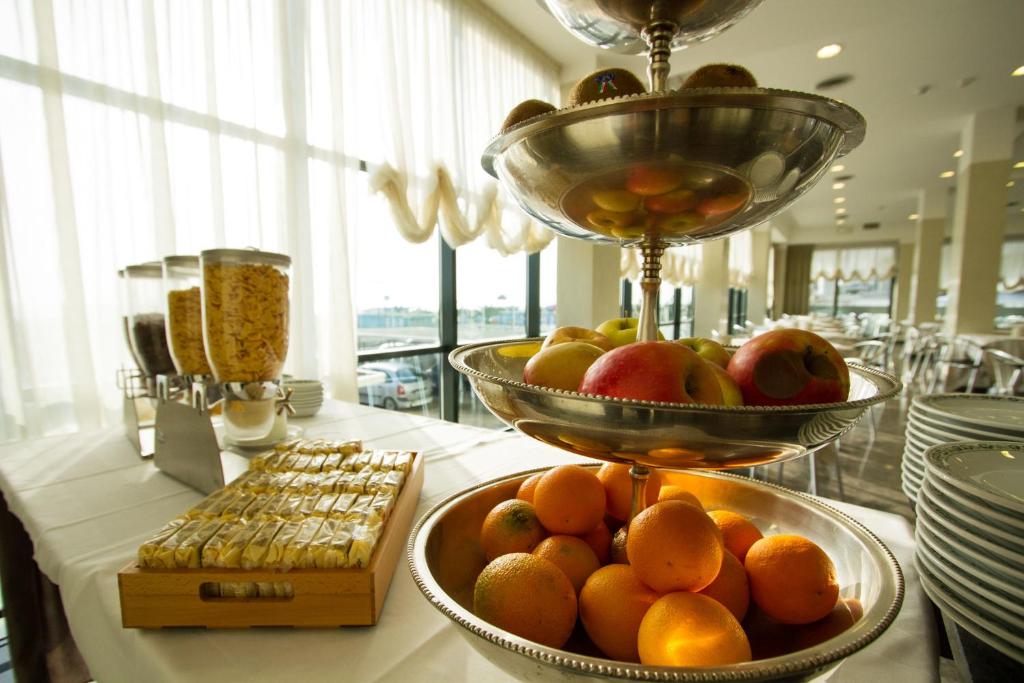Augļu stends ar dažādiem augļiem, tostarp apelsīniem un āboliem, atrodas viesnīcas brokastu zālē, kur ir arī graudaugi un saldumi. Gaisma ieplūst caur logiem, radot patīkamu atmosfēru.