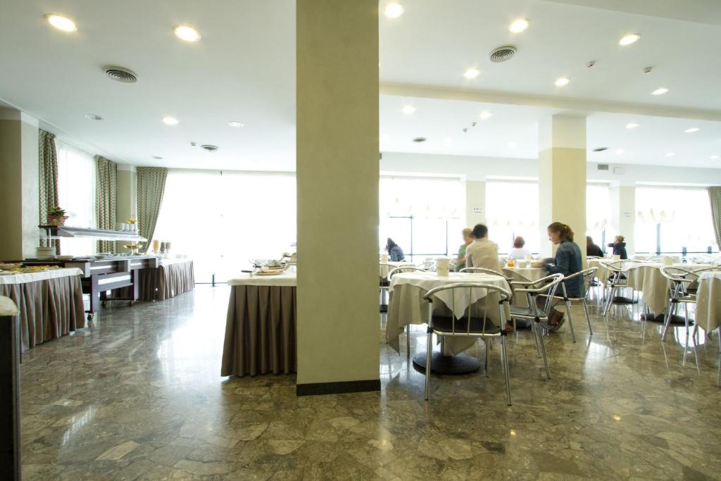 Gaiša restorāna iekārtojums ar elegantām galdniecībām un viesiem, kas baudījuši maltīti. Redzami bufetes galdi un plaša telpa ar dabīgu apgaismojumu.