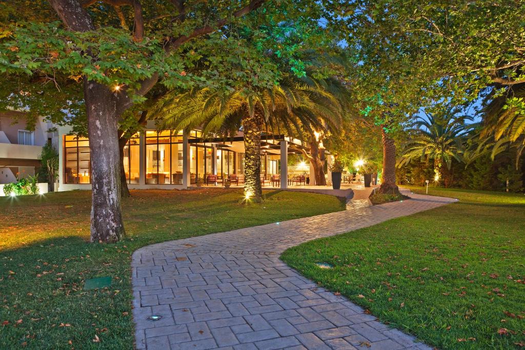 Skaists dārza ceļš, kas ved uz modernu ēku ar stikla fasādi, apņemts ar zaļām palmām un kokiem, radot mierīgu atmosfēru vakara gaismā.