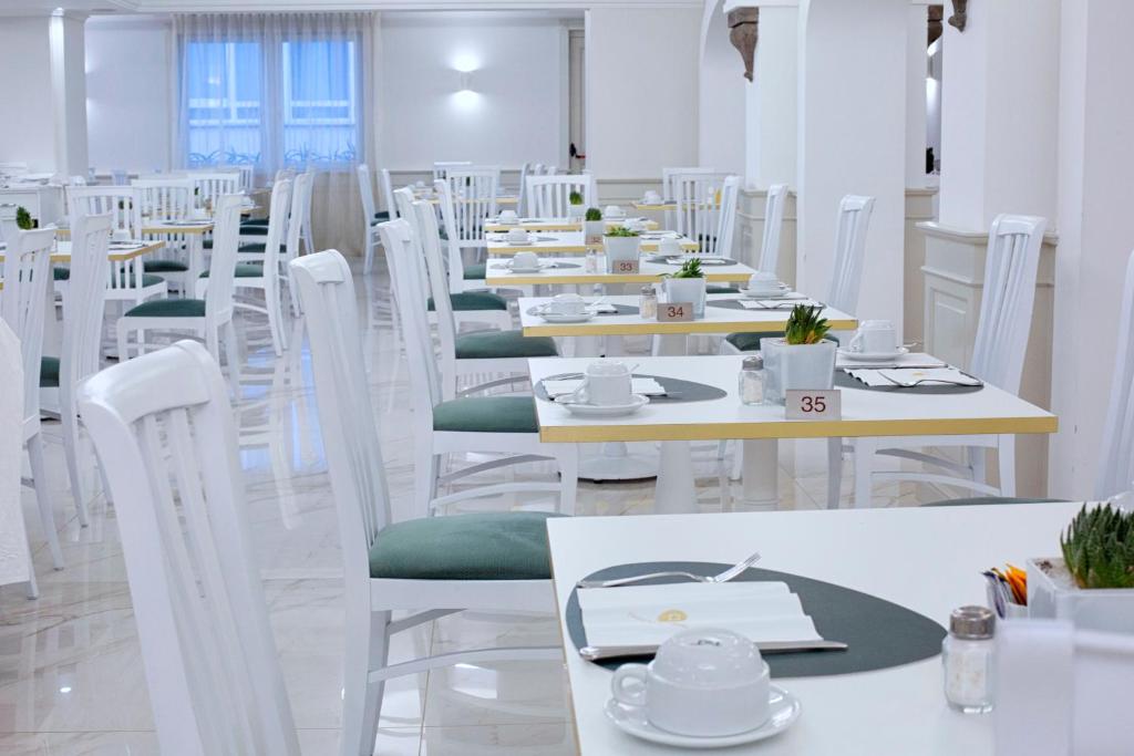 Gaišs un moderns restorāna interjers ar baltām galdiem un krēsliem, kas dekorēti ar zaļām detaļām. Uz galdiem ir trauki un augi, radot patīkamu atmosfēru.