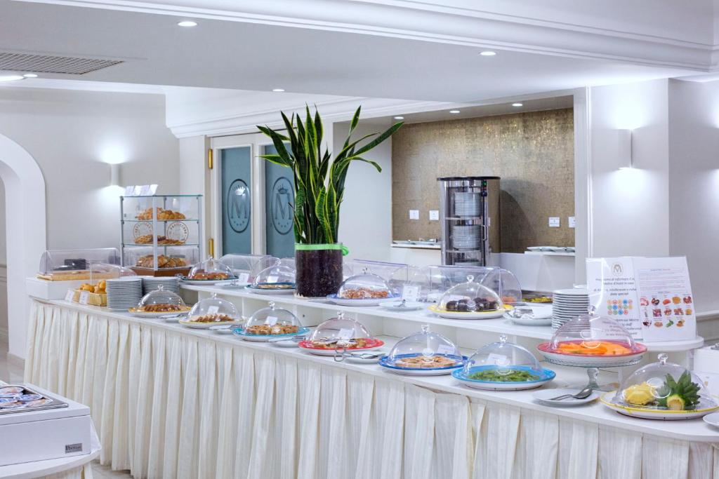 Bufete ar dažādiem ēdieniem un našķiem, kas pasniegti zem stikla vākiem, ar zaļiem augiem kā dekorāciju. Svaigi augļi un konditorejas izstrādājumi ir izkārtoti uz galda, radot pievilcīgu ēdiena piedāvājumu.