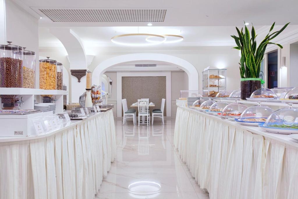 Gaismas un modernā dizaina bufete ar dažādiem saldumiem un uzkodām, izvietotiem zem stikla vākiem. Interjers ir gaišs un elegants, ar baltām galdautām un augiem.
