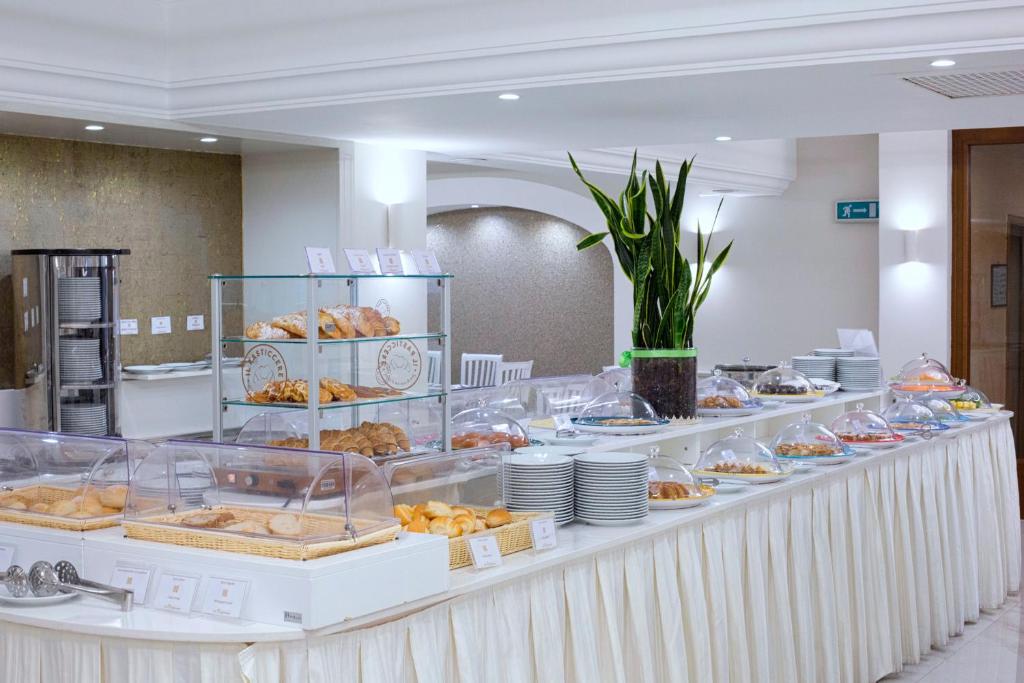 Bagātīga brokastu bufete ar dažādiem konditorejas izstrādājumiem un saldumiem, pasniegta gaišā un modernā vidē. Uz galda redzami arī šķīvji un dekoratīva auga kompozīcija.
