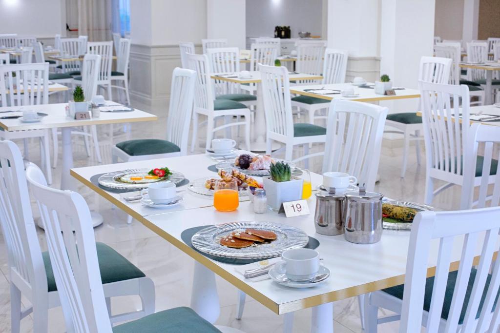 Gaiša un moderna brokastu zāle ar baltām galdiem un krēsliem, kur uz galda ir krāsu piesātināti ēdieni un dzērieni.