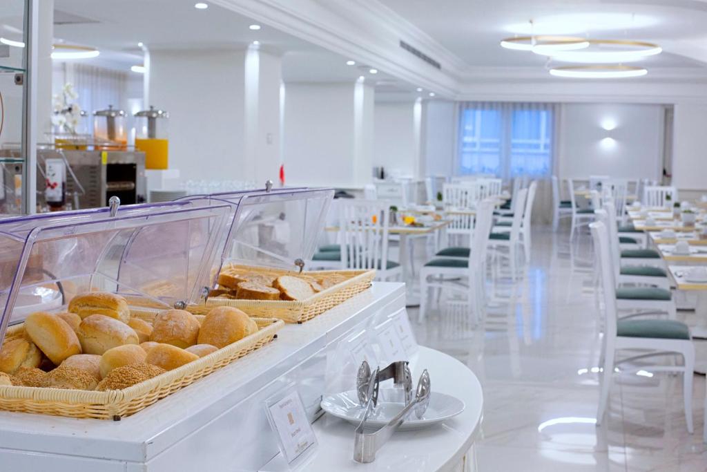 Elegants restorāns ar baltām mēbelēm un plašu ēdamzāli, kurā redzama maizes izvēle grozā uz bufetes galda.