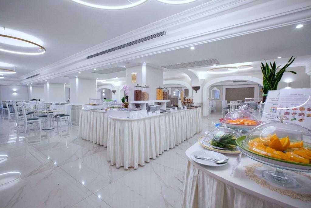 Mūsdienīga restorāna iekārtojums ar elegantām baltām mēbelēm un plašu bufeti, kas piedāvā dažādus ēdienus un augļus. Gaismas un interjers rada patīkamu atmosfēru.