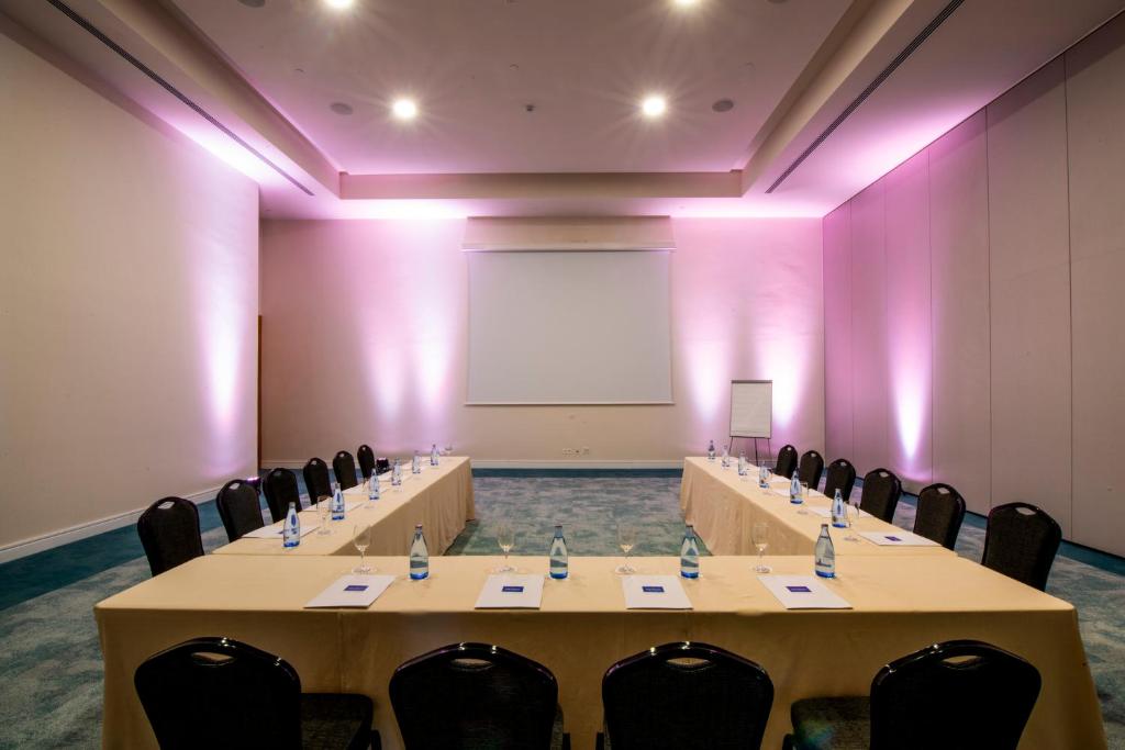 Konferenču telpa ar garu galdu, kas aprīkota ar ūdens pudelēm un papīra lapām, apgaismota ar rozā gaismu un lielu projektoru ekrānu.