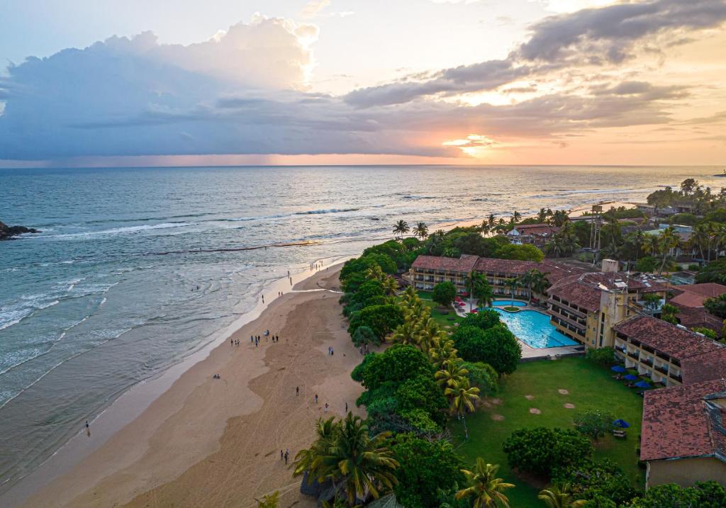 Pludmales aina ar saulrietu, kurā redzams viesnīcas komplekss ar baseinu un apzaļumotu teritoriju. Vairāki cilvēki pastaigājas pa pludmali, savukārt viļņi maigi sitas krastā.