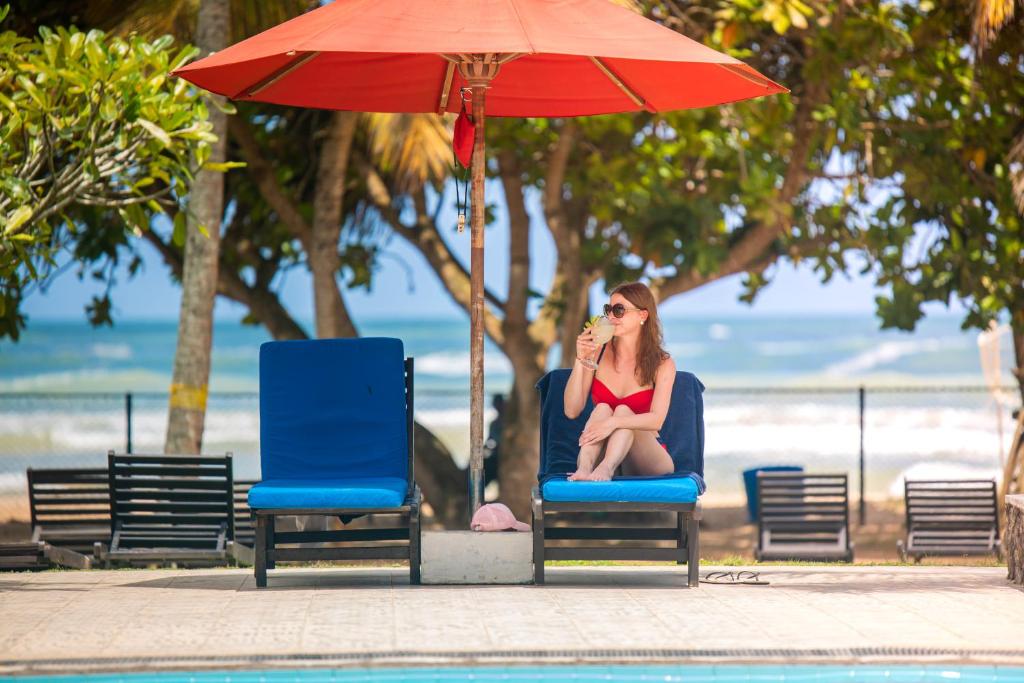 Sieviete sarkanos peldkostīmos atpūšas zem sarkanas saulessarga, turētā kokteiļa pie peldbaseina ar pludmales skatu.