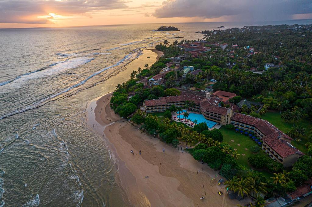 Pārskats uz pludmali ar viesnīcu kompleksu, palmām un okeānu, kur saulriets piešķir ainai siltas krāsas.