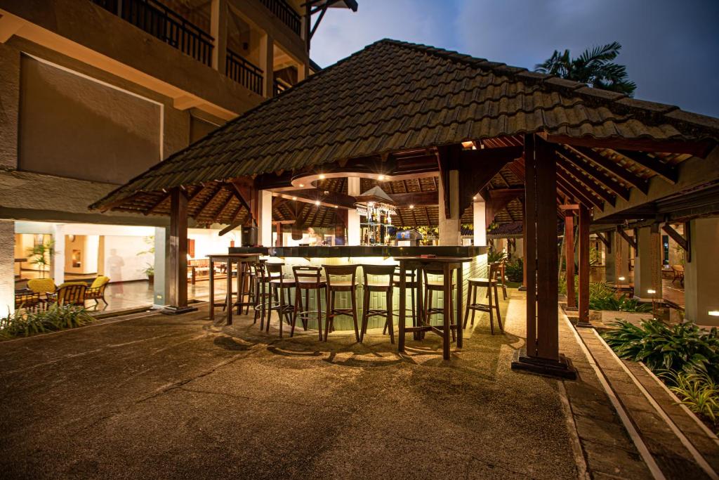 Āra bārs ar jumtu, apgaismots vakarā, ap kuru ir koka krēsli un zāle. Apkārt ir viesnīcas arhitektūra ar siltu apgaismojumu.