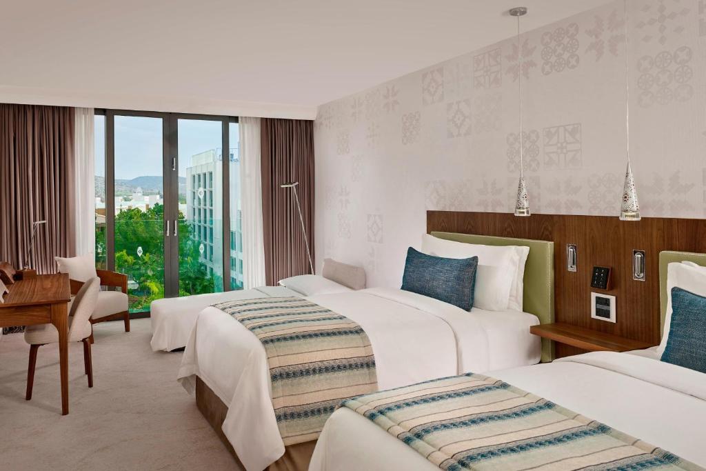 Mūsdienīga viesnīcas istaba ar divām gultām, koka mēbelēm un lieliem logiem, no kuriem paveras skats uz dārzu. Interjers ir gaišs un elegants, ar tekstilizstrādājumiem un mūsdienīgu apgaismojumu.