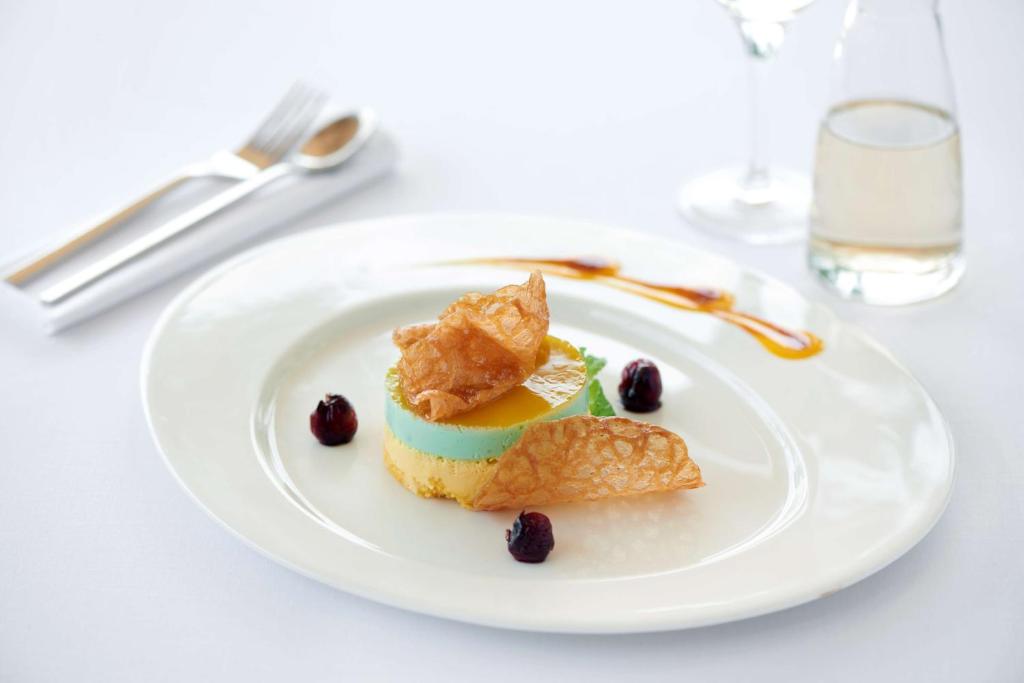 Krāsains desertu šķīvis ar trīs slāņiem: dzeltenu, zaļu un zilu, rotāts ar kraukšķīgu cepumu un ogām. Blakus glāze ar dzērienu un šķidruma trauks.