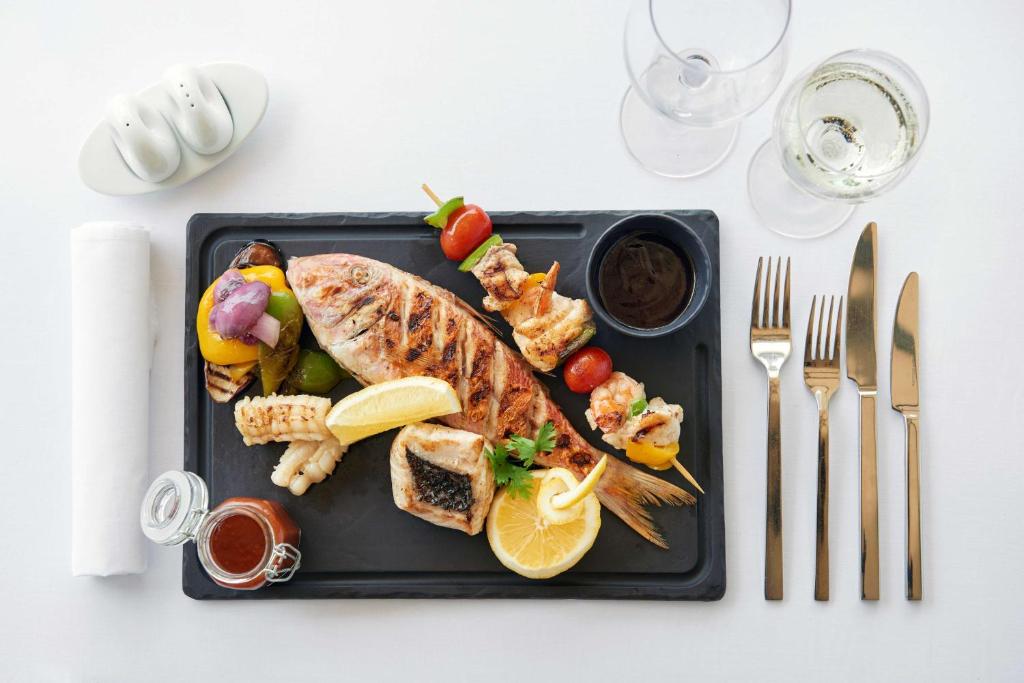 Grilēta zivs ar garnelēm, dārzeņiem un citrona šķēlēm, pasniegta uz melna šķīvja, blakus atrodas vīna glāze un piederumi.