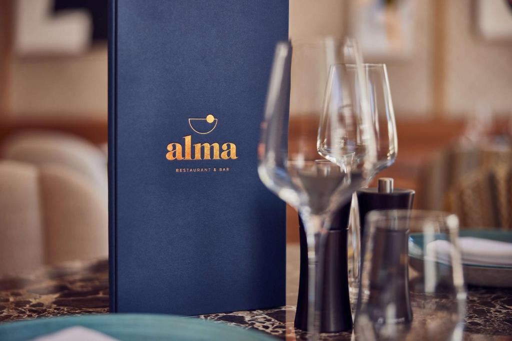 Restorāna "Alma" ēdienkarte uz elegantas galda virsmas ar glāzēm un garšvielu traukiem.
