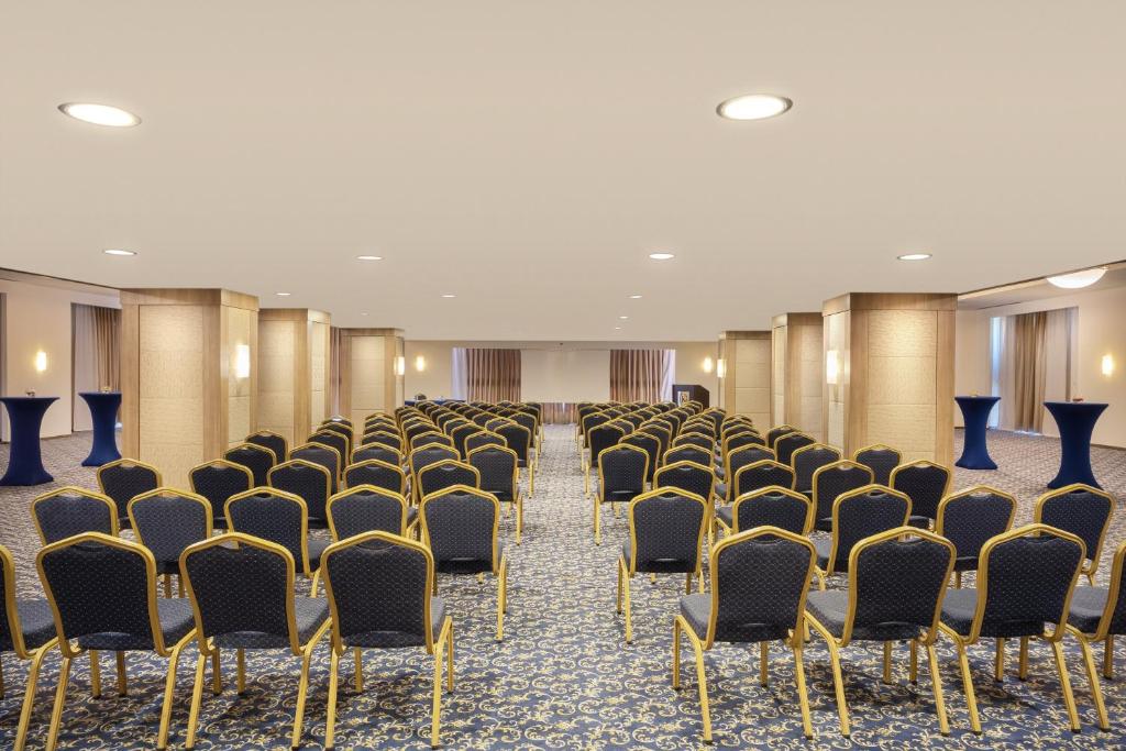 Konferenču zāle ar rindu sēdvietām un elegantu interjeru, gatava pasākumiem. Telpā ir redzami zili galdi un logi ar aizkariem.