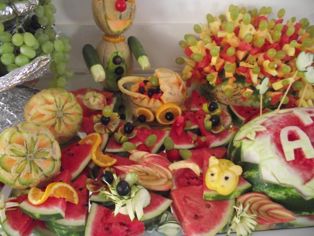Krāšņs augļu un dārzeņu izkārtojums, kas veidots no arbūziem, meloniem un vīnogām, radot jautrus figūriņas un dekorācijas. Šis radošais ēdienu noformējums ir ideāls svinībām vai svētku pasākumiem.