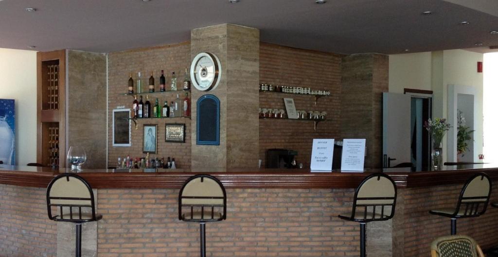 Bārs ar koka letēm un akmens sienu, uz kuras izvietoti dzērienu pudeles un aksesuāri. Gaisma ir maiga, radot relaksējošu atmosfēru.