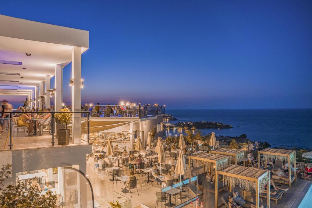 Panorama Village Hotel restorāna terase vakara krēslā ar viesiem un skatu uz jūras piekrasti.