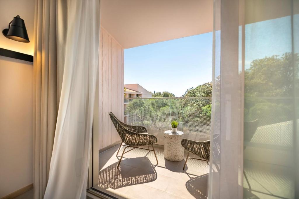 Balkona skats ar divām modernām krēslām un nelielu kafijas galdiņu, apzaļumotā apkārtnē. Gaisma ieplūst caur caurspīdīgām aizkariem, radot mierīgu atmosfēru.