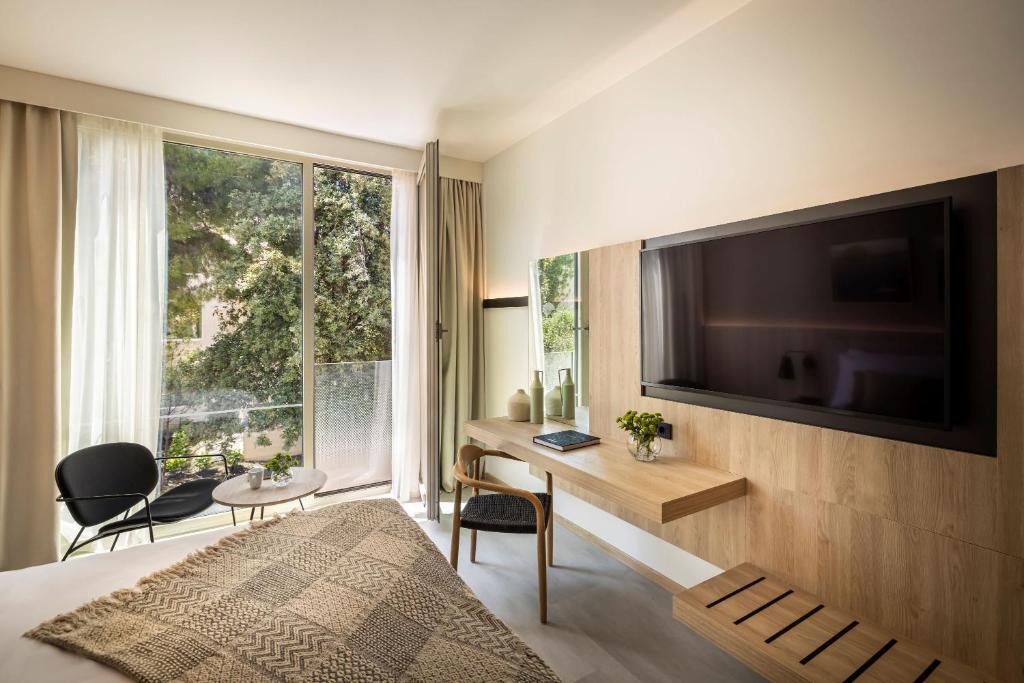 Mūsdienīga viesnīcas numura interjers ar plašu logu, kas paver skatu uz dārzu, un elegantu mēbelējumu, tostarp gultu, TV un galdu ar krēslu.