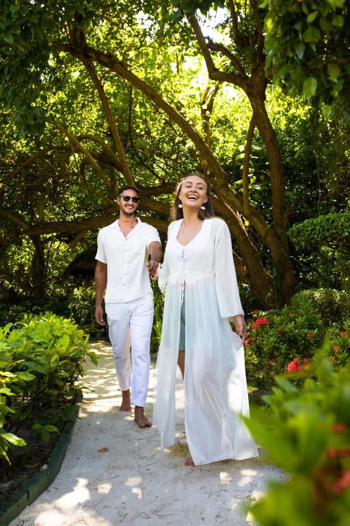 Pāris pastaigā pa saulainu dārzu, tērpti vieglos, vasarīgos apģērbos, izstaro prieku un mīlestību. Apzaļumotā vide un ziedi veido idillisku fonu romantiskai atmosfērai.