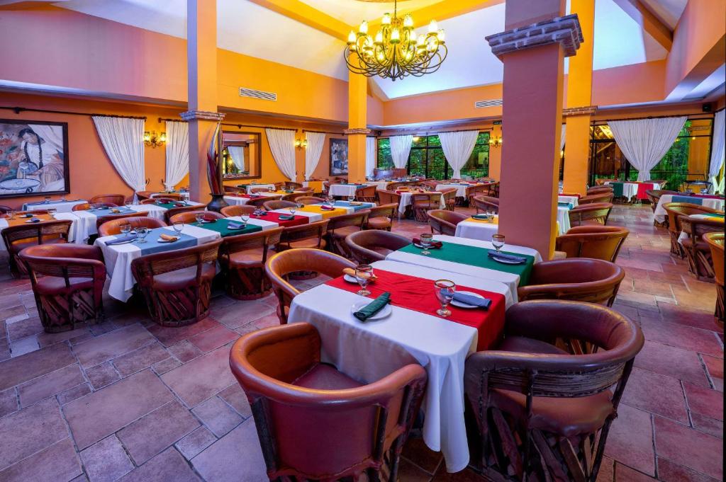 Restorāna iekšskats ar krāsainiem galdiem, kas klāti ar baltām un zaļām galdautiem, un koka krēsliem. Telpa ir apgaismota ar piekaramo lampu un rotāta ar mākslas darbiem uz sienām.