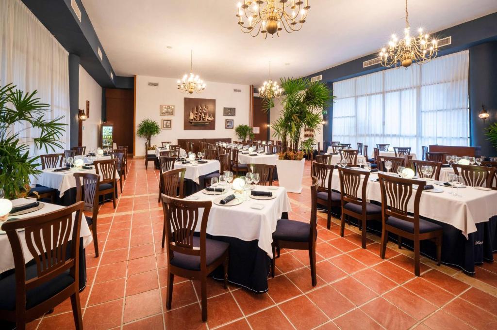 Elegants restorāna interjers ar gaišām krāsām, koka mēbelēm un dekoratīviem augiem. Galds ir klāts ar baltām galdautām un galda piederumiem, radot patīkamu atmosfēru.