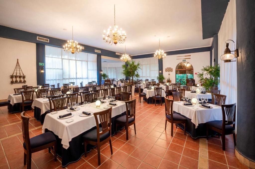 Eleganti iekārtota restorāna telpa ar baltām galdautām un koka mēbelēm, kas piedāvā patīkamu atmosfēru. Gaismas un zaļumi rada mājīgu noskaņu.