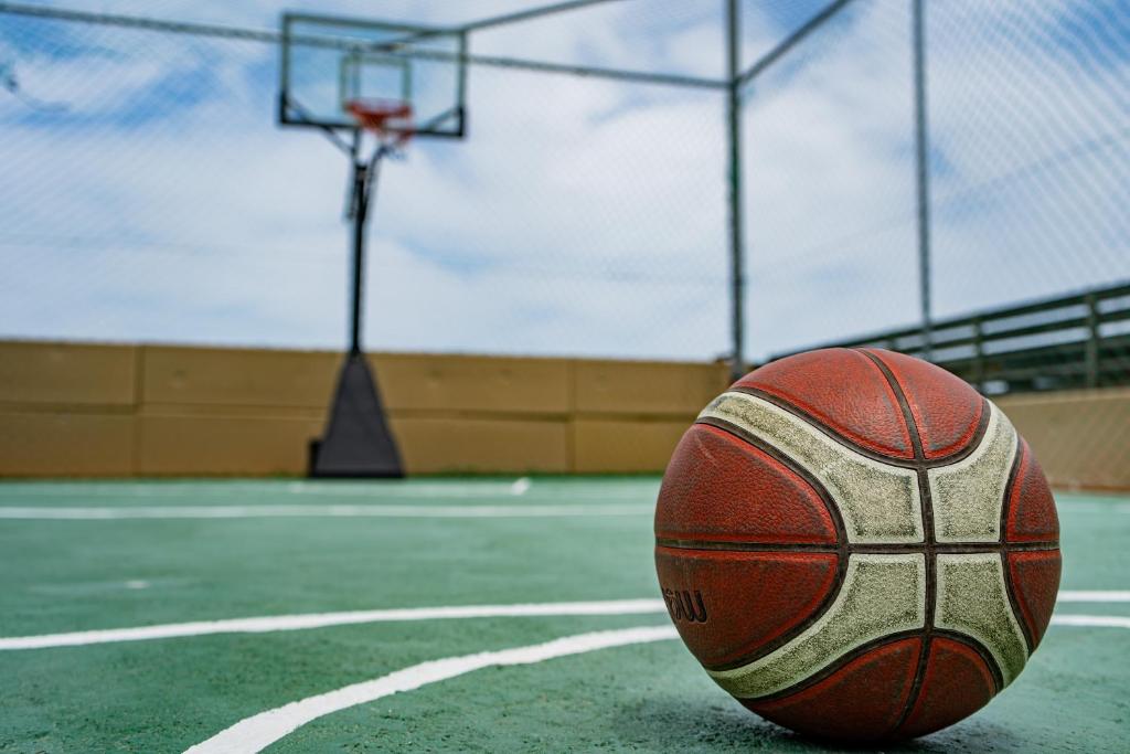 Basketbola bumba uz zaļā laukuma ar grozu fonā, skaidra debesis un sporta laukuma elementi.