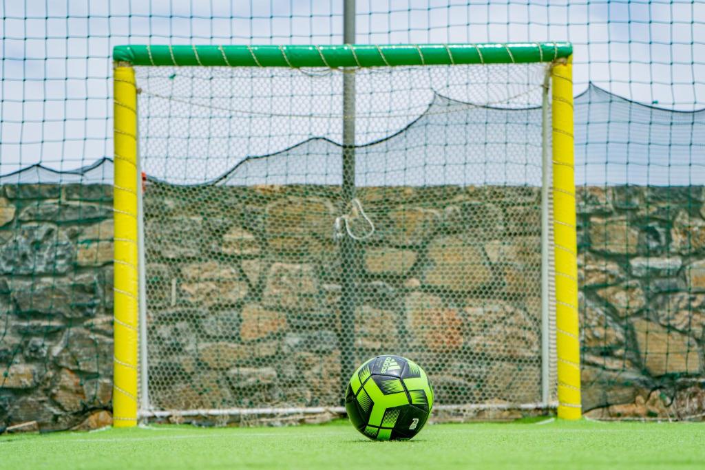 Futbola vārti ar zaļu un dzeltenu apmalīti, priekšplānā melna un zaļa bumba uz mākslīgā zāliena.