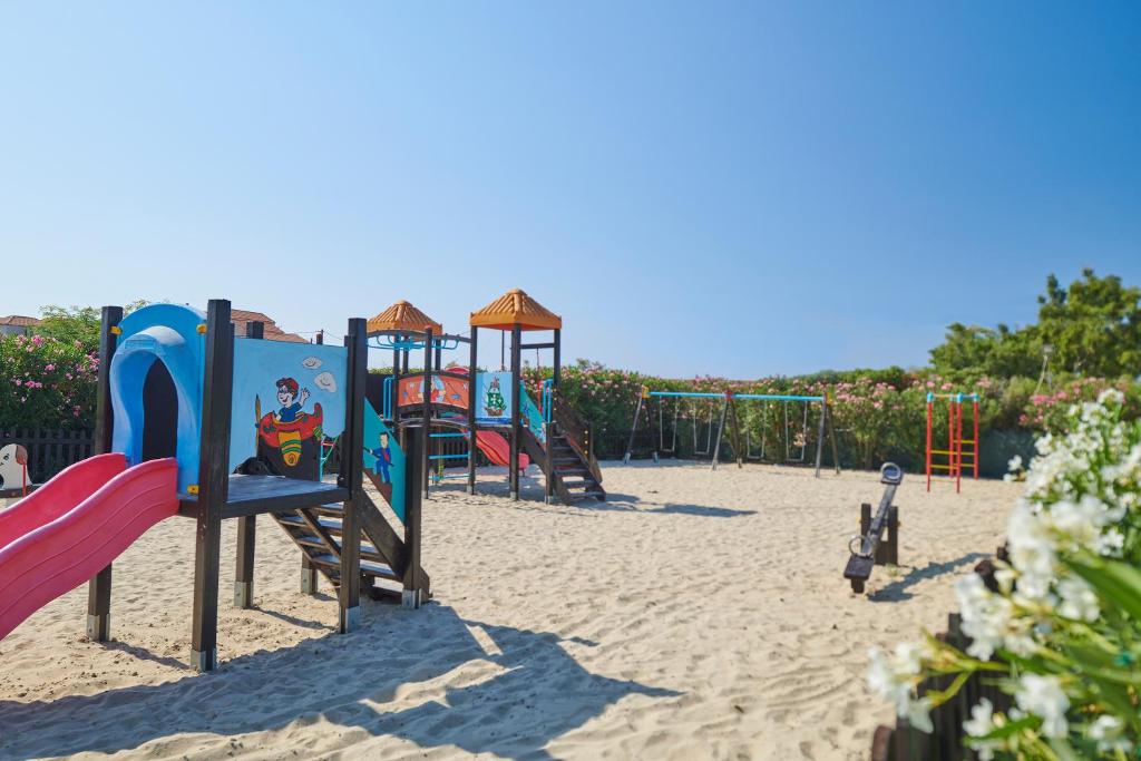 Bērnu rotaļu laukums ar slidkalniņu, šūpolēm un krāsainām rotaļām, ieskauts ziedu un zaļumu ainavā. Ideāla vieta bērniem, lai spēlētos un izbaudītu saulainu laiku.