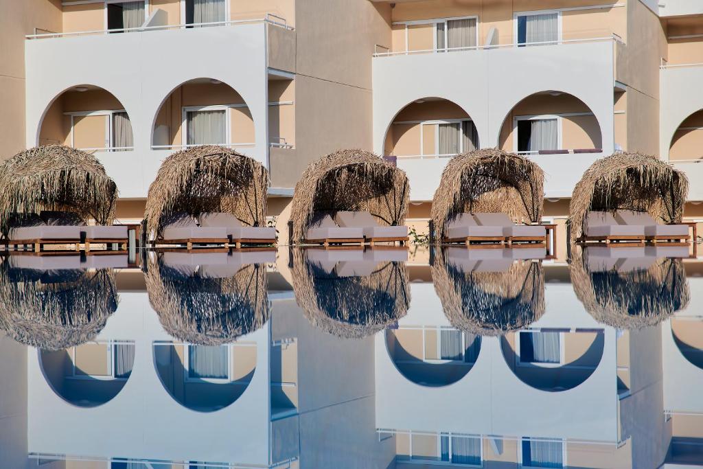 Lieliskas peldvietas arēnas ar salmu nojumeēm, kas atspoguļojas ūdenī, radot mierīgu atmosfēru. Fons veido elegantas viesnīcas ararkas.