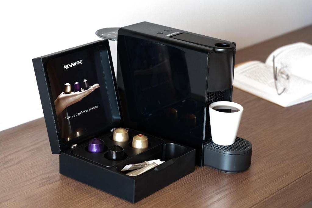 Nespresso kafijas automāts ar kapsulām un krūzi uz koka galda, blakus atvērtai grāmatai un brillēm. Ideāls risinājums kafijas baudīšanai mājās.