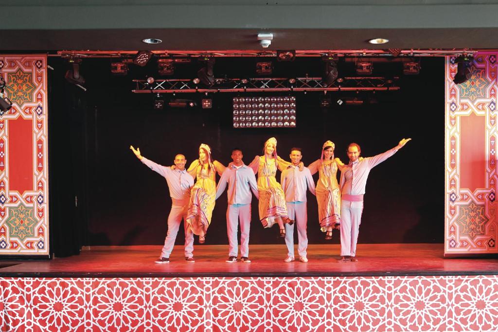 Dejotāju grupa uz skatuves, tērpta krāsainās tradicionālās drēbēs, izpilda deju izrādi, fona dekorācijas veido krāsainu dizainu.