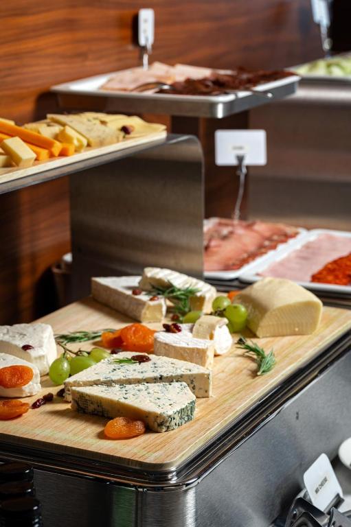 Siera šķīvja ar dažādām sieru šķirnēm, žāvētiem augļiem un vīnogām, kas izkārtoti uz koka griešanas dēļa. Fonā redzami citi pārtikas produkti, piemēram, gaļa un dārzeņi.