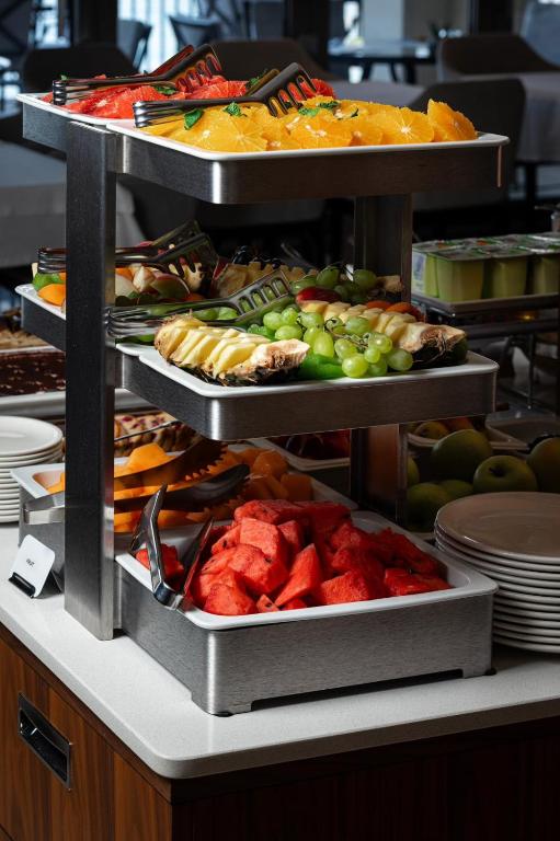 Augļu bufete ar dažādiem svaigiem augļiem, tostarp arbūzu, ananāsu, vīnogām un apelsīniem, izkārtota trīs stāvos. Galdautā redzami arī paplātes un šķīvji, kas pieejami apmeklētājiem.