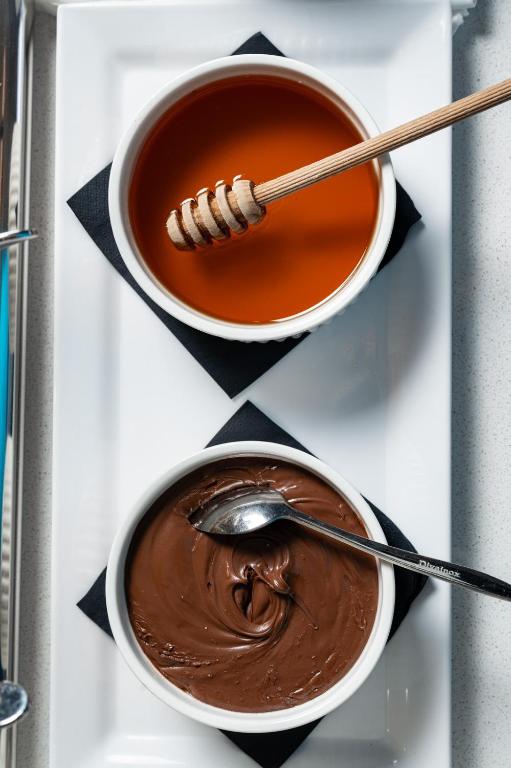 Divas bļodas ar medu un šokolādes krēmu, pasniegtas uz balta šķīvja ar melnu salveti. Medus bļodā ir koka karote, savukārt šokolādes krēmā ir metāla karote.