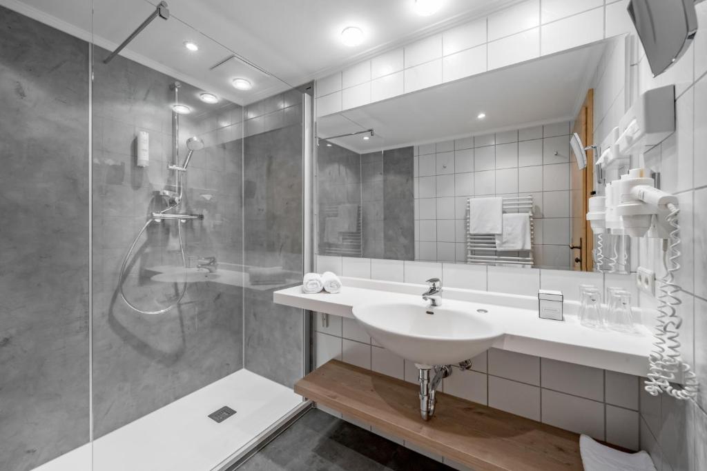 Mūsdienīga vannas istaba ar stikla dušu, baltu izlietni un koka plauktu, nodrošinot elegantu un funkcionālu dizainu. Gaismas un flīžu kombinācija rada patīkamu atmosfēru.