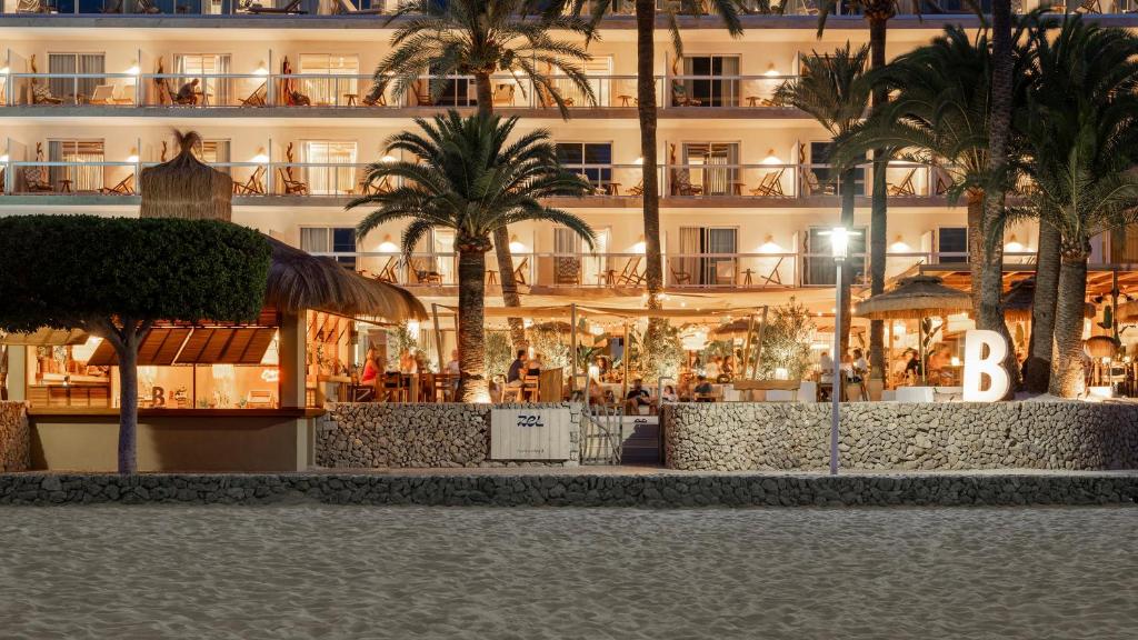 Pludmales restorāns ar palmām un apgaismotām terasēm, kas atrodas blakus viesnīcai. Vakarā redzams, kā cilvēki bauda maltītes un atpūtu.
