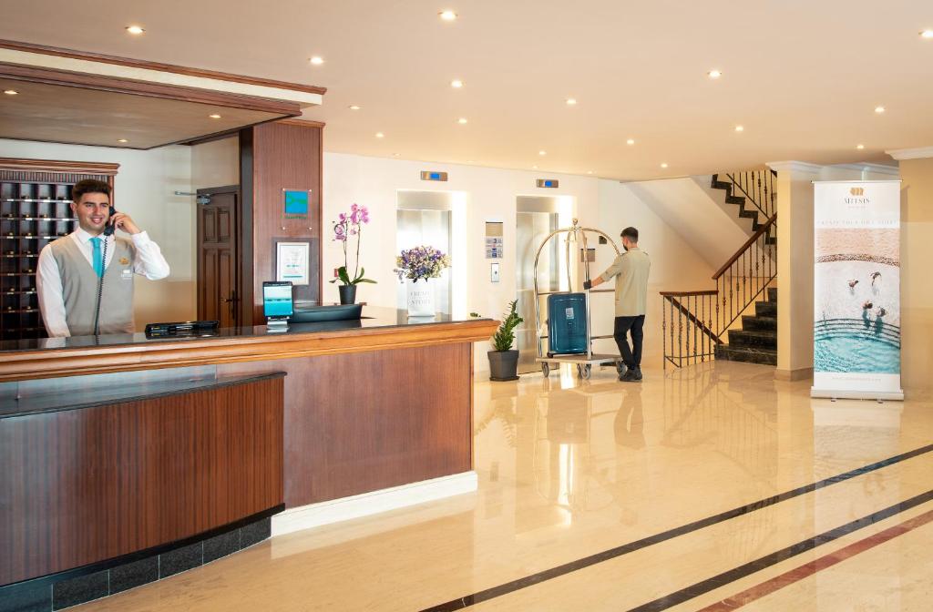 Reģistratūra viesnīcā ar laipnu darbinieku, kas runā pa telefonu, un viesi, kas pārvadā bagāžu. Gaismas un moderna iekārtojuma atmosfēra.