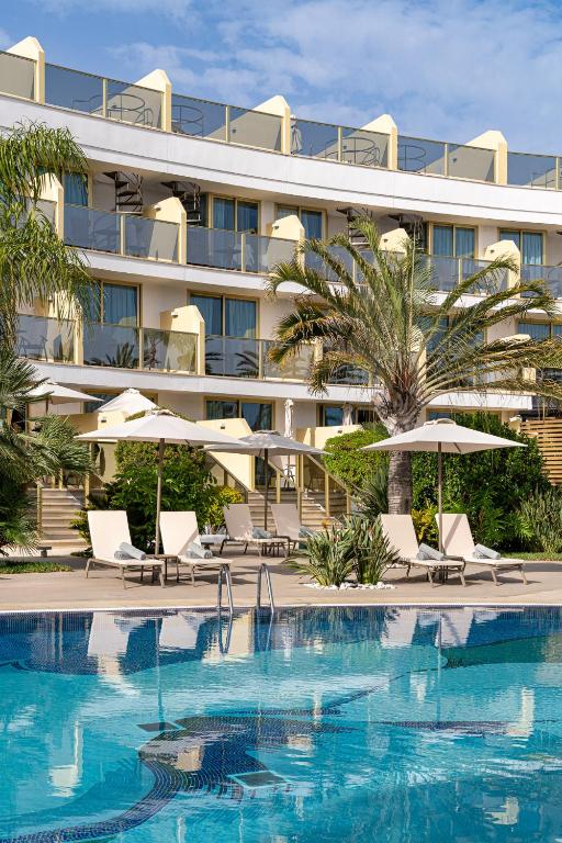 Luksusīgs viesnīcas komplekss ar baseinu, saules gultām un palmām, kas rada atpūtas atmosfēru. Skats uz ēku ar balkoniem un zilo debesi.