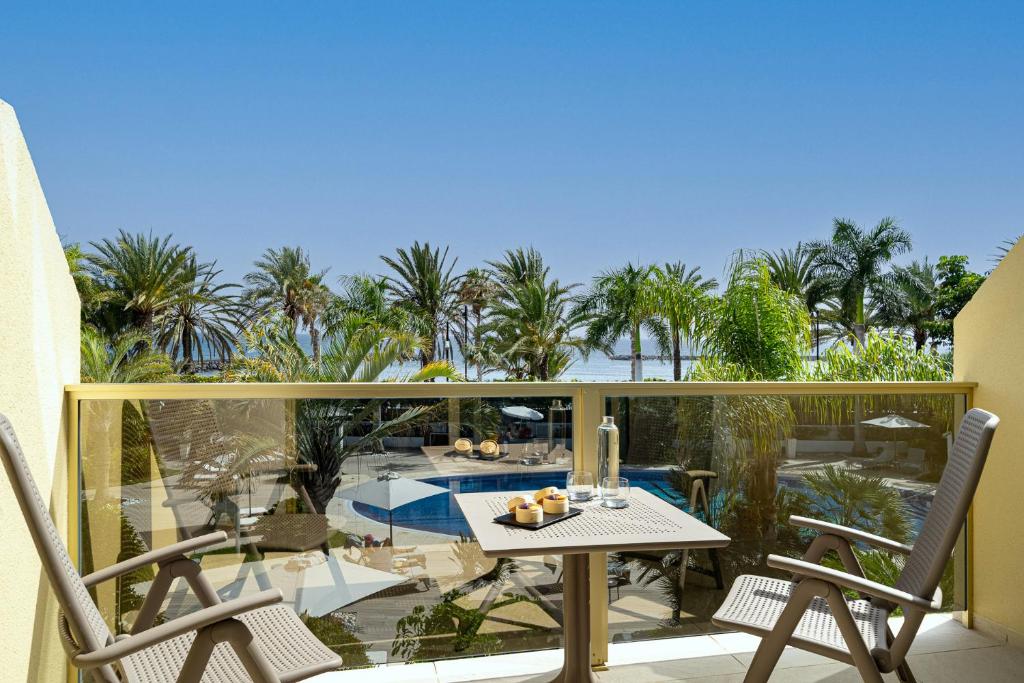 Balkona skats uz baseinu un palmām, kas nodrošina mierīgu un relaksējošu atmosfēru. Uz galda ir ūdens pudele un deserti, kas papildina atpūtas mirkļus.