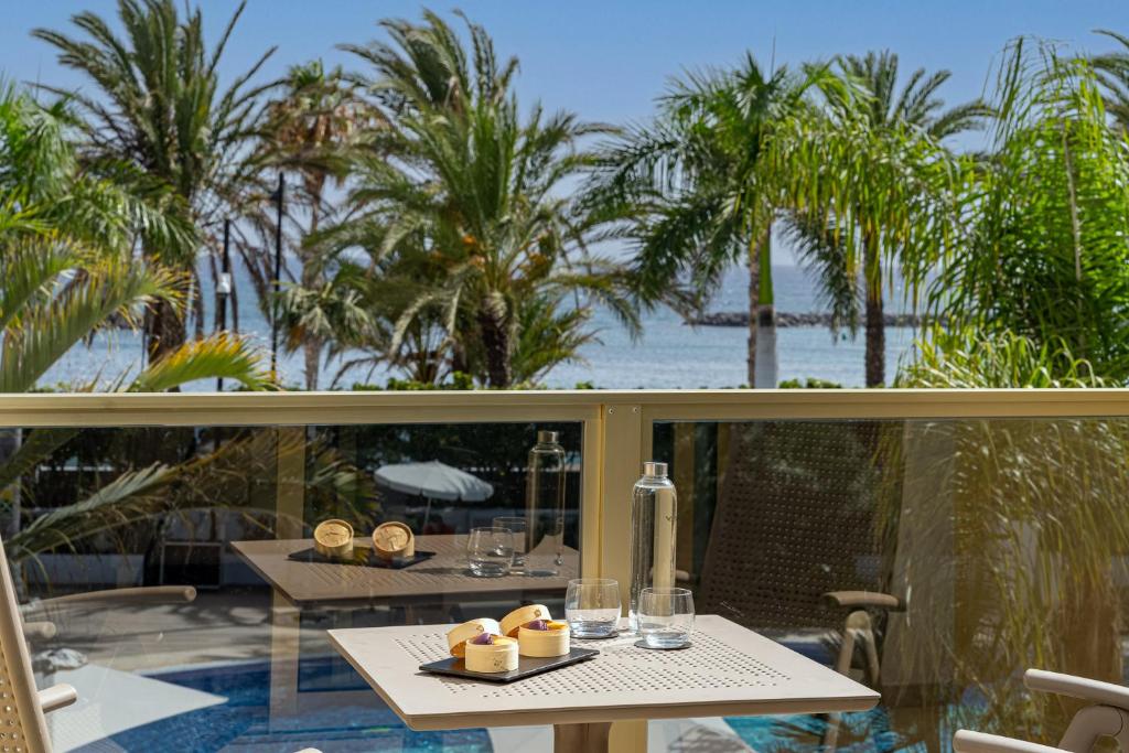 Skats no balkona uz jūru, kur uz galda ir deserts un ūdens, ar palmām fonā. Ideāla vieta relaksācijai un baudīšanai.